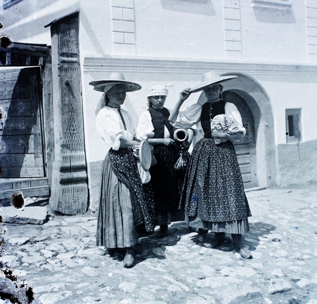 Torockói asszonyok és lányok ünnepi viseletben 1907-ben: fehér főkötő, hímzett kötény, csizma, a háttérben a falu házai